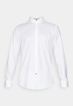 Chemise blanche à manches longues en tissu lisse, avec un col classique, une fermeture à boutons sur le devant, et un ourlet arrondi avec un petit détail de bouton accentué.