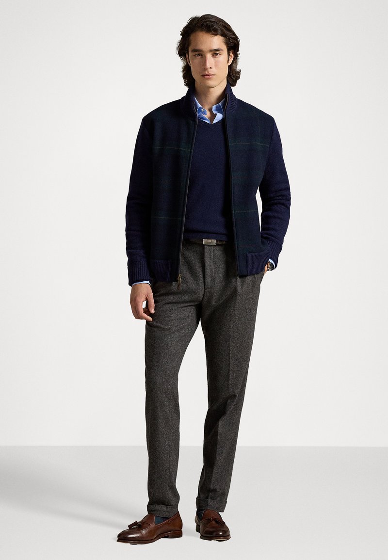 Navy zip-up jacket with plaid pattern, dark sweater, light blue shirt, gray trousers, and brown leather shoes. Textured materials and tailored fit.