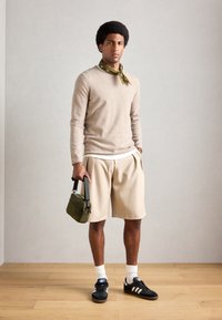 Beige Pullover, grün gemusterter Schal, cremefarbene, gefaltete Shorts, schwarze Sneaker mit weißen Streifen und grüne Tasche mit Schultergurt.