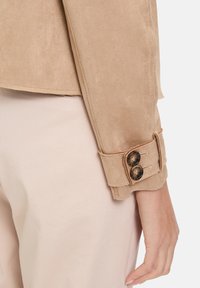 Manche d'une veste en daim beige avec une fermeture à double bouton, présentant des boutons texturés marron, exposée à côté d'un pantalon de couleur claire.