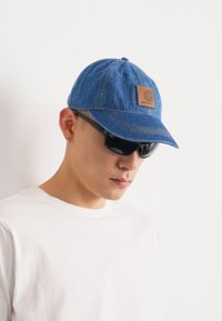 Blå denimcaps med en brun læderlogo-patch, parret med sorte solbriller. Skjorten er ensfarvet hvid, og baggrunden er neutral.