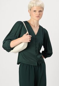 Mørkegrønn bluse med V-hals og knappedetaljer, med puffermer. Kombinert med matchende bukser og en teksturert hvit skulderveske.