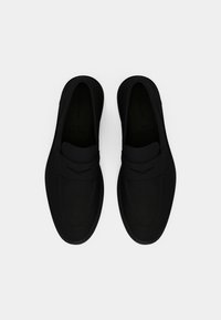 Ett par svarta slip-on loafers med en snygg design, runda tår och en bred remdetalj över toppen på en vit bakgrund.