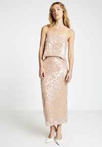 Roségoldfarbenes, paillettenbesetztes Kleid mit figurbetonter Silhouette, verstellbaren Spaghettiträgern und langem Rock. Kombiniert mit spitzen weißen Absätzen.