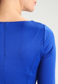 Robe ajustée bleue à manches longues, présentant une texture lisse, un col rond et un détail discret de fermeture éclair au dos.