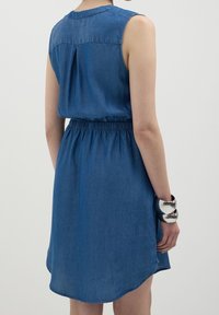 Hihaton farkkumekko keskisinisessä, jossa on kokoontaitettu vyötärö, sileä pinta ja kaareva helma. Hopeinen rannekoru korostaa rannetta.