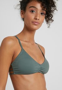 Zeleni trikotni bikini top z tankimi naramnicami, ki vsebuje zasnovo z nabranim materialom in stranskimi izrezi. Gladka tekstura, lahek material.