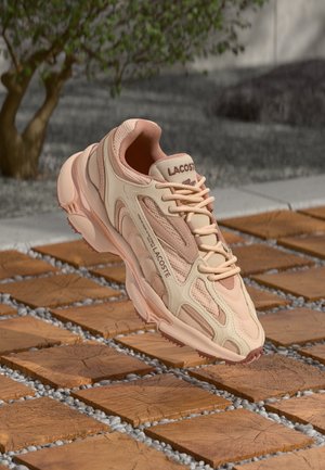 Baskets Lacoste beige et rose flottant au-dessus d'un bloc de bois et d'un chemin de galets avec un feuillage vert flou en arrière-plan.