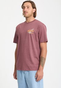 Bordeauxrotes Baumwoll-T-Shirt mit kurzen Ärmeln, das ein gelbes grafisches Logo auf der linken Brust hat. Lockere Passform mit Rundhalsausschnitt.