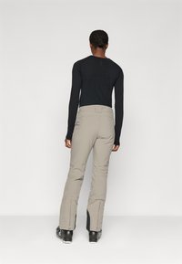 Beige smučarske hlače s ozko obliko, z zadrgo na spodnjem delu nogavic in prilegajočim pasom, v kombinaciji z dolgimi črnimi rokavi.