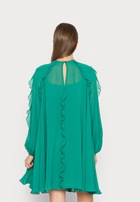 Vestido verde de chiffon com mangas longas e com babados, costas com abertura em forma de chave e silhueta fluida. Textura suave e detalhes de babados em camadas ao longo das bordas.