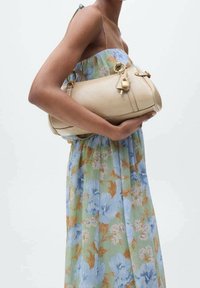 Beige Lederhandtasche mit goldfarbener Hardware und einem Schloss, unter dem Arm gehalten, vor einem blumigen Kleid in Grün-, Blau- und Orangetönen.