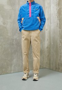 Pullover in pile blu con patta a bottoni rosa e tasche con zip arancioni; abbinato a pantaloni cargo beige e sneakers beige.