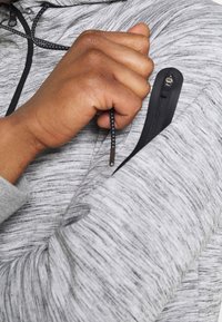 Gray, textured pullover with a zippered arm pocket; features black accents and a drawstring. Close-up of hand reaching into the pocket.