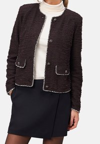 Veste noire texturée avec garniture argentée, col rond, boutons et poches avant. Portée sur un col roulé blanc et une jupe.