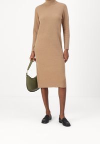 Robe en maille beige avec un col montant et des manches longues, associée à un sac à main vert et des mocassins noirs. Texture lisse et coupe mi-longue.