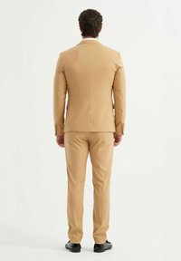 Veste et pantalon couleur chair, coupe ajustée, tissu texturé, vue de dos, fente simple, avec des chaussures noires. Les boutons accentués sont visibles.