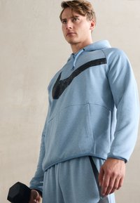 Világoskék kapucnis pulóver, puha anyagból, fekete swoosh logóval a mellkasán. A modell egy fekete súlyzót tart a kezében, bemutatva az edzőruhákat.