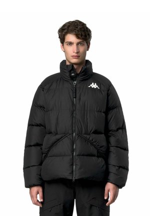 Giovane con i capelli ricci che indossa una giacca imbottita nera con una cerniera frontale e un logo bianco sul petto, in piedi davanti a uno sfondo bianco.