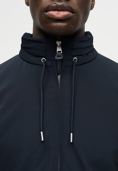 Κοντινό πλάνο ενός ατόμου που φοράει σκούρο μπουφάν με φερμουάρ, ψηλό γιακά και κορδόνια που κρέμονται μπροστά.