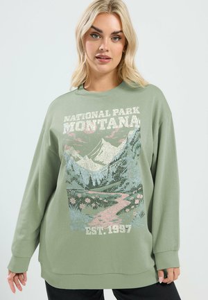 Blonde Frau trägt übergroßen salbeigrünen Pullover mit Montana Nationalpark malerischem Druck und Text "Est. 1937".