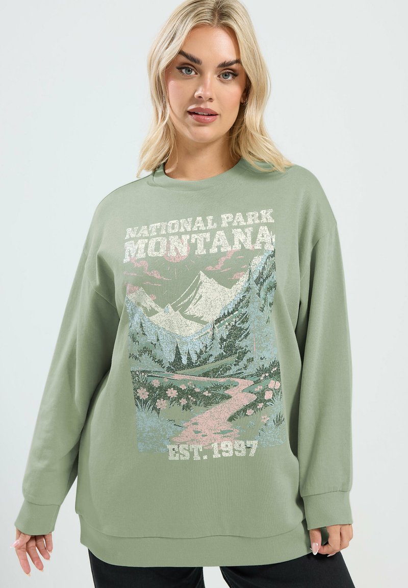 Blonde Frau trägt übergroßen salbeigrünen Pullover mit Montana Nationalpark malerischem Druck und Text "Est. 1937".