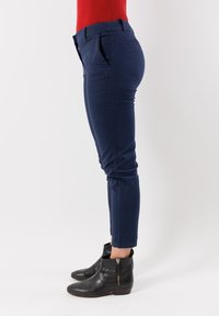 Persona che indossa pantaloni blu scuro aderenti, una maglietta rossa e stivaletti neri con cerniere laterali, in piedi di lato su uno sfondo bianco semplice.