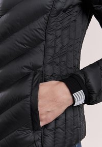Veste en duvet noire matelassée avec un motif diagonal, poches latérales et poignets côtelés avec un accent rayé. Matériau lisse et brillant.