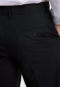 Schwarze Anzughose mit flachem Bund, Seitentaschen und einer einzigen Gesäßtasche, die mit einem schwarzen Knopf gesichert ist. Glatte Stoffoberfläche.