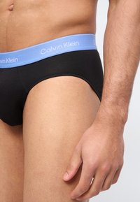 Κοντινό πλάνο ενός ατόμου που φορά μαύρα εσώρουχα με ανοιχτό μπλε ζωνάκι Calvin Klein, εμφανίζοντας το κάτω μέρος του κορμού και το δεξί χέρι σε χαλαρή στάση.