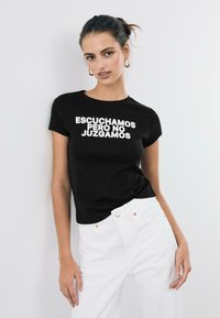 Schwarzes Baumwoll-T-Shirt mit weißem Text "ESCUCHAMOS PERO NO JUZGAMOS." Kombiniert mit hoch taillierten weißen Hosen, ohne sichtbare Muster oder Verzierungen.