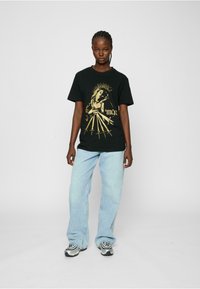 Black cotton t-shirt featuring gold graphic print of a figure with rays and swords, paired with light blue wide-leg jeans and sneakers.