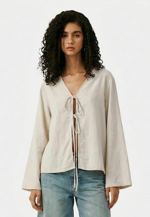 Žena s kučeravými vlasmi má na sebe voľné béžové tričko zaväzované dvakrát vpredu a svetlomodré džínsy, stojí pred jednofarebným pozadím.