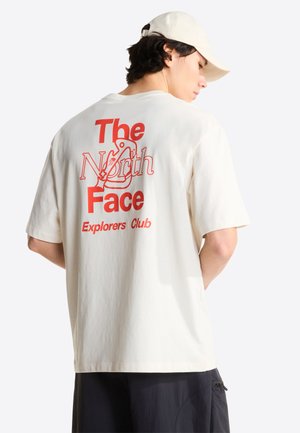 Mężczyzna w kremowej czapce i białej koszulce z czerwonym napisem i grafiką "The North Face Explorers Club" na plecach, czarne spodnie, stoi bokiem.