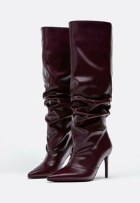 Burgundské čižmy po kolená vyrobené z lesklej kože, s špicatou špičkou a vysokým ihlovým podpätkom, so zberaným detailom na holenici.