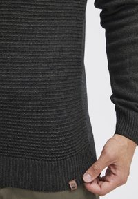 Pulover tricotat de culoare gri închis, cu un model texturat, tiv drept și o eticheta mică cu logo aproape de margine. Material moale la vedere.