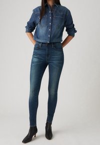 Camisa de mezclilla oscura con dos bolsillos en el pecho y cierre de botones, combinada con vaqueros ajustados de talle alto en azul oscuro y botas negras hasta el tobillo.