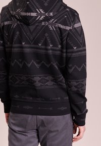 Svart sweatshirt med huva, geometriska mönster i gråtoner samt ribbade muddar och nederkant. Tyget ser mjukt ut med en texturerad design.