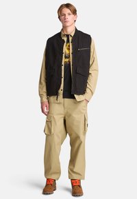 Zwarte stoffen vest met voorzakken, gedragen over een beige langtermijn shirt en een zwart T-shirt met opdruk, gecombineerd met beige cargobroek en bruine laarzen.