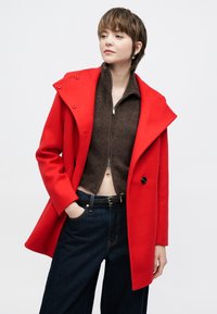 Red, tailored coat with a wide collar and double-breasted design. Worn over a brown zip-up sweater and dark denim pants.