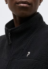 Κοντινό πλάνο ατόμου που φοράει μαύρο υφασμάτινο φλις μπουφάν με ψηλό γιακά και λευκό κεντημένο λογότυπο στο στήθος.