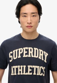 Camiseta de algodón azul marino con letras de chenille color crema que dicen "SUPERDRY ATHLETICS", manga corta y cuello redondo.