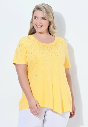Frau lächelt, trägt ein gelbes Kurzarmshirt mit kleinen Verzierungen und weiße Hose, steht vor weißem Hintergrund.