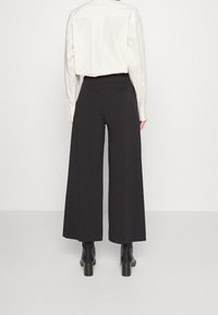 Zwarte wijde broek met een hoge taille. De stof heeft een gladde textuur en is gedetailleerd met opvallende verticale naden. Gecombineerd met zwarte laarzen met hak.