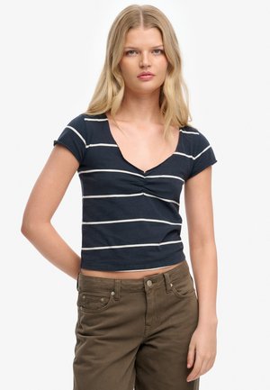 Young woman with long blonde hair wearing a navy blue cropped T-shirt with white stripes and high-waisted brown pants.