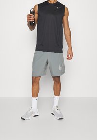 Czarna sportowa koszulka bez rękawów, jasnoszare krótkie spodenki z logo, białe skarpetki i szare sneakersy. Trzyma czarną butelkę na wodę.