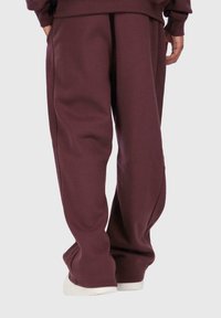 Person in lockeren, maroonfarbigen Sweatpants und weißen Schuhen, stehend mit entspannten Händen an den Seiten vor einem schlichten Hintergrund.