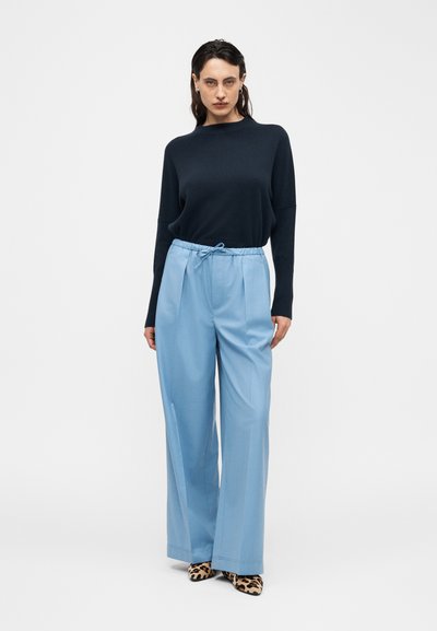 Femme debout portant un pull à manches longues noir, un pantalon large bleu clair avec une taille à cordon de serrage, et des chaussures pointues à imprimé léopard.