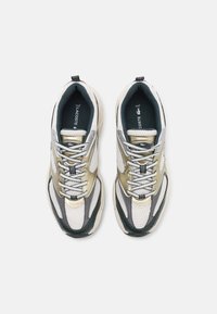 Buty sportowe Lacoste mają siateczkową i syntetyczną cholewkę w kolorach białym, srebrnym i ciemnozielonym, miękką podeszwę i płaskie sznurowadła.