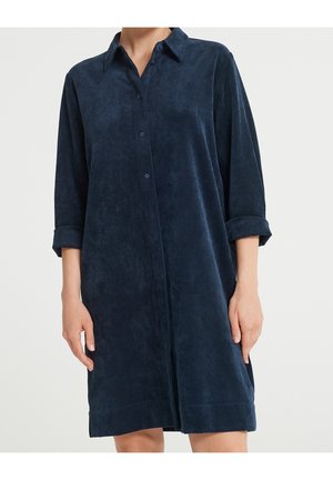 Donna che indossa un vestito camicia blu navy, a maniche lunghe con bottoni e maniche arrotolate, di lunghezza al ginocchio, su sfondo neutro.